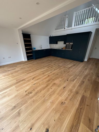 Spacious room with wooden flooring, featuring a dark kitchen area and high ceilings.